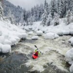 Bracing For The Coming PFAS Ban a person paddles down a river in a snowy winter landscape while wearing a drysuit that respects the upcoming PFAS ban