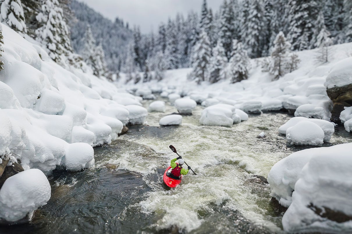 a person paddles down a river in a snowy winter landscape while wearing a drysuit that respects the upcoming PFAS ban