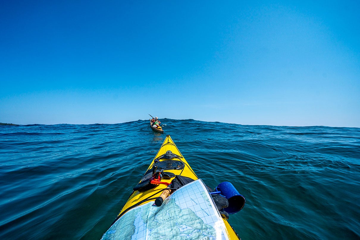 bow shot of a sea kayaker with map paddling after another kayaker over an ocean swell while they are on an expedition