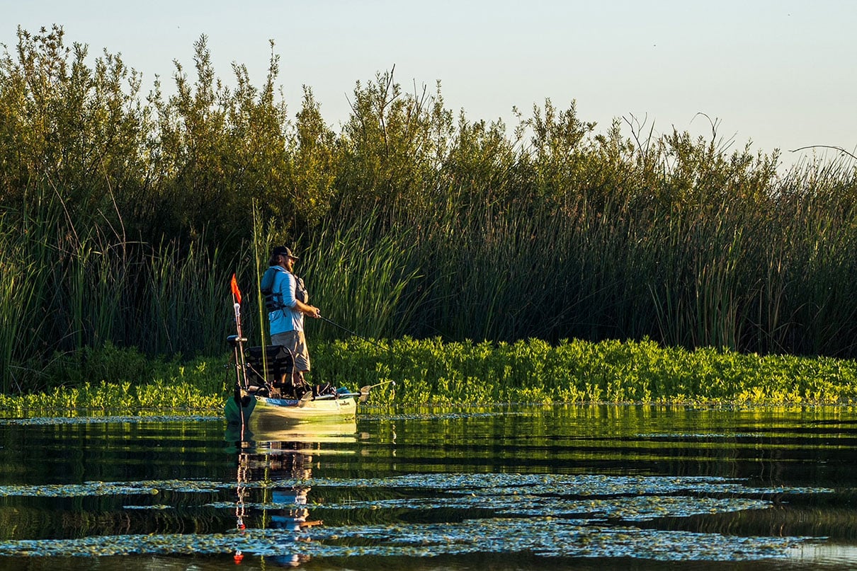 man stands and fishes from a kayak near grass flats in golden afternoon light