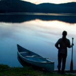 person stands at dawn or dusk holding a canoe paddle beside a canoe parked at the shore of a calm waterway with hills behind