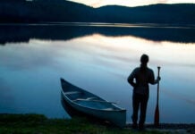 person stands at dawn or dusk holding a canoe paddle beside a canoe parked at the shore of a calm waterway with hills behind