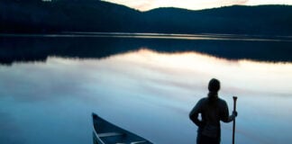 person stands at dawn or dusk holding a canoe paddle beside a canoe parked at the shore of a calm waterway with hills behind