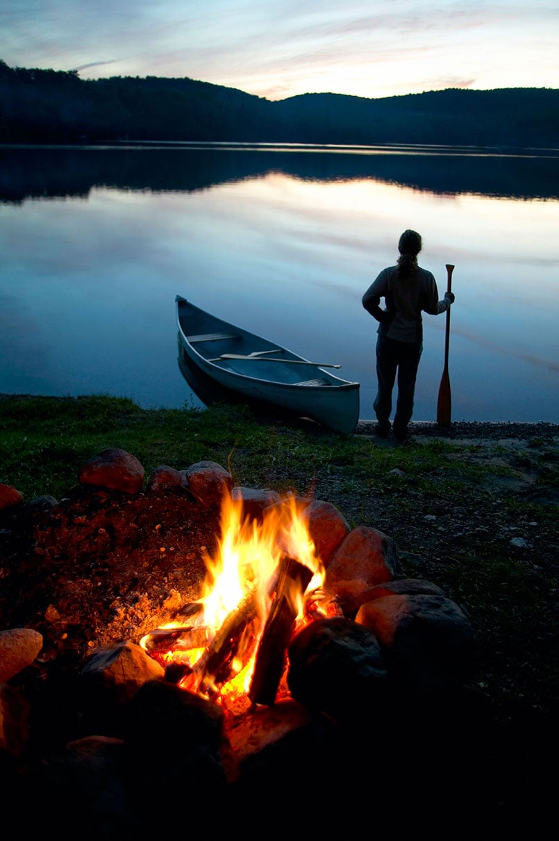 person stands at dawn or dusk holding a canoe paddle beside a canoe parked at the shore of a calm waterway with hills behind