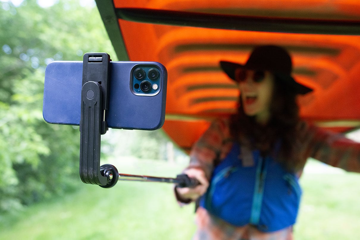 woman smiles at her phone held on a selfie stick while portaging a canoe