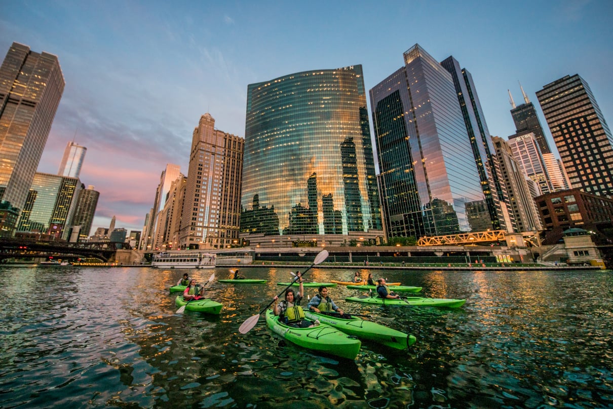 Group in kayaks below city skyline at sunset.